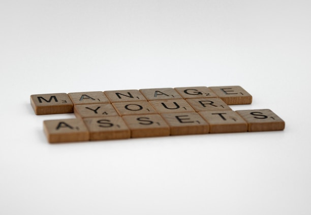 Wooden Scrabble tiles arranged in an organized pattern spell out the phrase 'MANAGE YOUR ASSETS' on a plain white background.