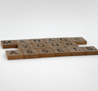 Wooden Scrabble tiles arranged in an organized pattern spell out the phrase 'MANAGE YOUR ASSETS' on a plain white background.