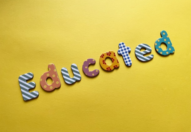 Colorful letters spelling the word 'Educated' are arranged on a bright yellow background. Each letter features a different pattern, such as stripes, polka dots, flowers, and stars.