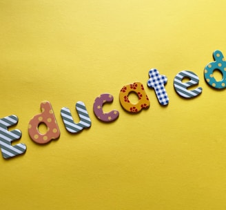 Colorful letters spelling the word 'Educated' are arranged on a bright yellow background. Each letter features a different pattern, such as stripes, polka dots, flowers, and stars.