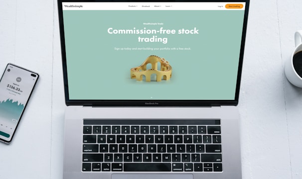 A laptop with a financial website open is centered on a white table. The screen displays a mint green background with text related to commission-free stock trading, accompanied by a gold object resembling a bridge. To the left, a smartphone displays a financial app with a graph and monetary figure. A cup of coffee sits on the right side, adding a casual work environment atmosphere.