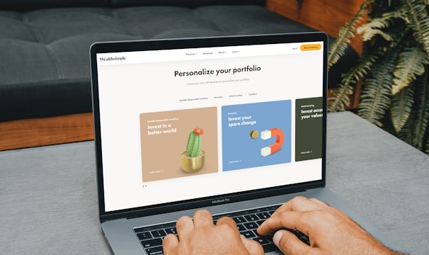 A person is typing on a MacBook Pro, viewing a website titled 'Personalize your portfolio.' The webpage displays three sections related to investing, each with different images and text. The laptop is on a grey table with a dark sofa and indoor plants in the background.