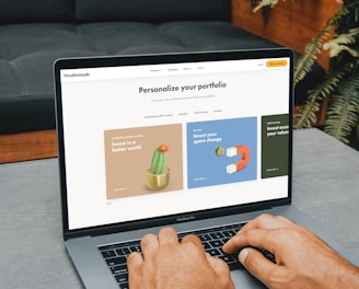 A person is typing on a MacBook Pro, viewing a website titled 'Personalize your portfolio.' The webpage displays three sections related to investing, each with different images and text. The laptop is on a grey table with a dark sofa and indoor plants in the background.