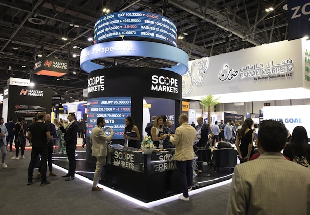 A bustling exhibition hall filled with people attending a financial conference or trade event. Prominent displays and booths from companies like Scope Markets, Daman Markets, and Hantec Markets are visible. Attendees are engaged in conversations, networking, and exploring the offerings and information provided by the exhibitors.