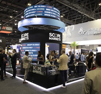 A bustling exhibition hall filled with people attending a financial conference or trade event. Prominent displays and booths from companies like Scope Markets, Daman Markets, and Hantec Markets are visible. Attendees are engaged in conversations, networking, and exploring the offerings and information provided by the exhibitors.