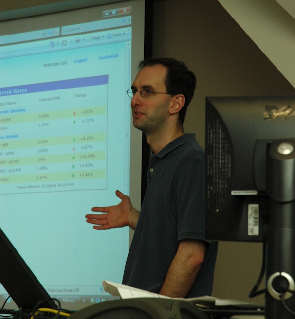 A person standing in front of a projector screen displaying interest rates and other financial information. The person appears to be giving a presentation or lecture in a classroom or office setting. A computer monitor and some papers are visible on a desk in the foreground.