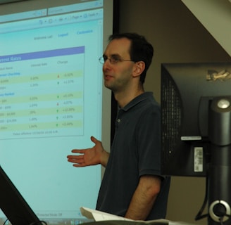 A person standing in front of a projector screen displaying interest rates and other financial information. The person appears to be giving a presentation or lecture in a classroom or office setting. A computer monitor and some papers are visible on a desk in the foreground.