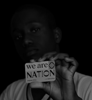 A person holds a card close to the camera, with a focused, serious expression. The card reads 'we are NATION Empower Africa' with a dark background that contrasts with the white text and card. The lighting highlights the card more than the person's face.