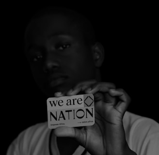 A person holds a card close to the camera, with a focused, serious expression. The card reads 'we are NATION Empower Africa' with a dark background that contrasts with the white text and card. The lighting highlights the card more than the person's face.