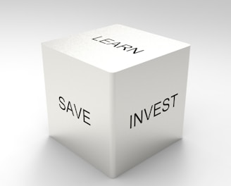 A three-dimensional cube with words on its visible sides, featuring 'LEARN' on the top, 'SAVE' on the left face, and 'INVEST' on the right face. The cube is rendered in a minimalist white and gray color palette, creating a clean and professional look.